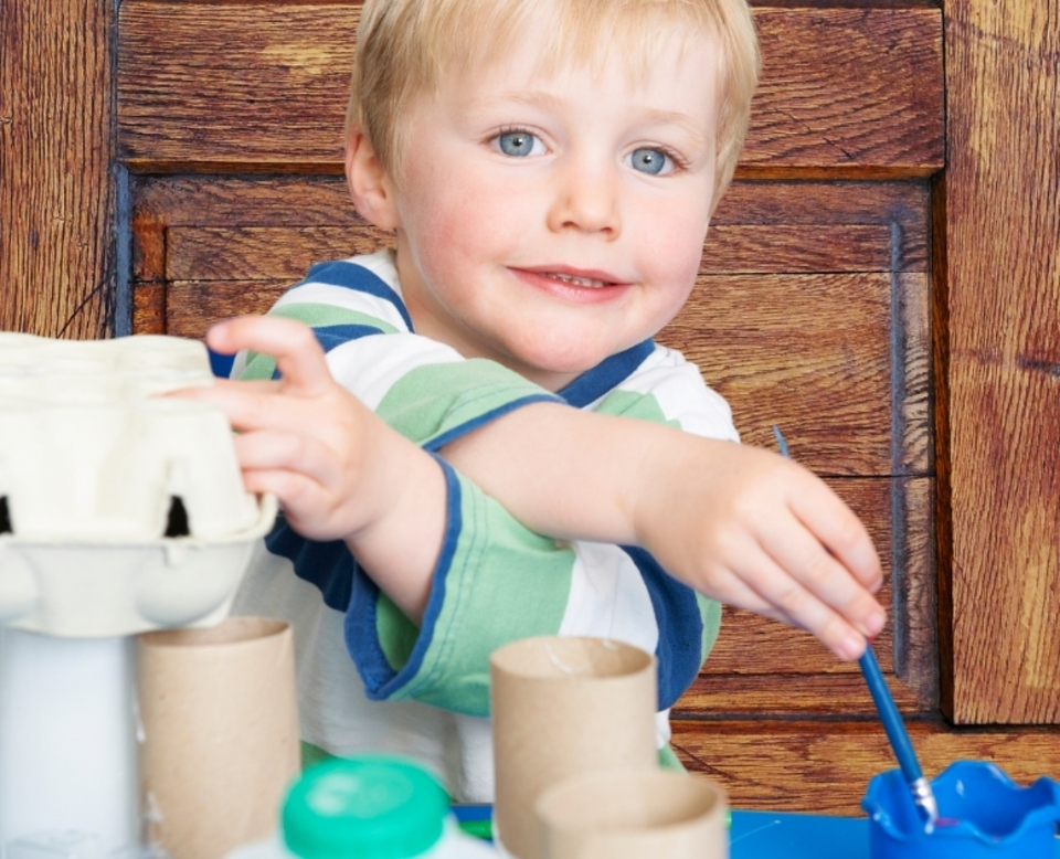 A young child with blond hair smiles while painting and playing with recycled craft materials, such as cardboard tubes and egg cartons, at a blue table in front of a wooden background.